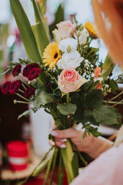 A person holding a vibrant bouquet of fresh flowers featuring soft pink roses, white freesias, bright yellow gerbera daisies, and deep red carnations, arranged with greenery and wrapped with clear cel
