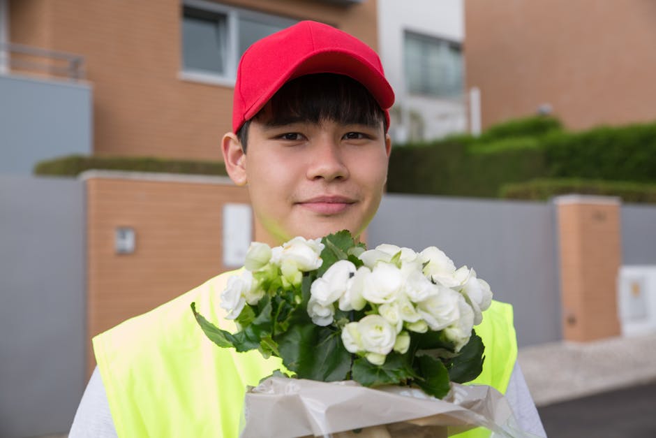 A young male flower delivery courier wearing a bright yellow safety vest and red cap, standing outdoors in front of modern residential buildings with a neatly trimmed lawn. He holds a bouquet of fresh