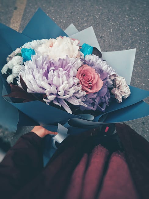 A hand holding a bouquet of fresh flowers wrapped in blue and light blue paper, featuring a large white chrysanthemum, a soft pink rose, smaller purple and white blossoms, and bright blue accents. The