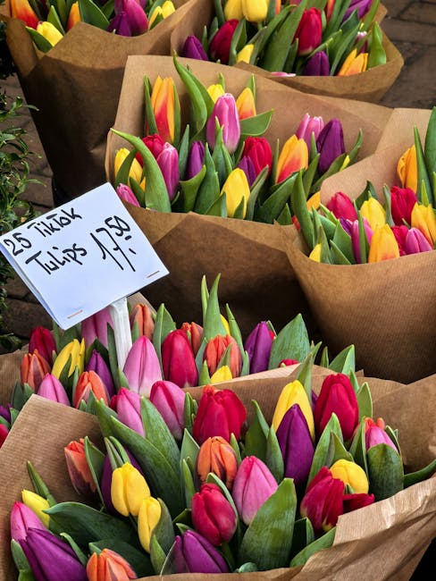 A collection of fresh tulip bouquets in various colors including pink, purple, yellow, orange, and red, each wrapped in brown craft paper. The tulips are arranged with green leaves and sturdy stems, a