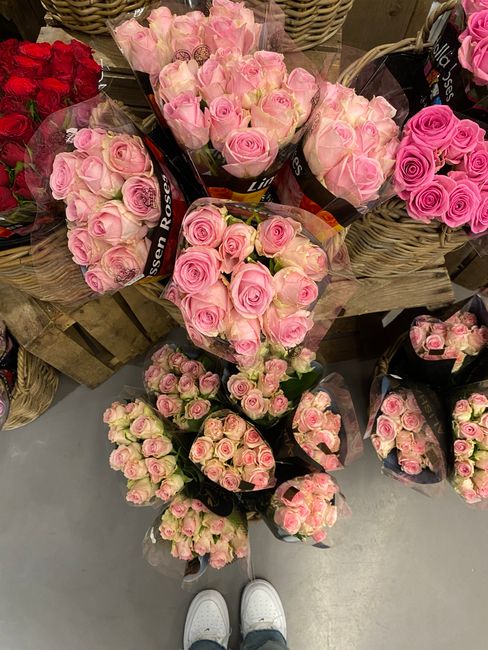 A collection of fresh pink roses arranged in bouquets and smaller clusters, featuring delicate, tightly packed petals and green stems. The flowers are wrapped in clear and black floral paper, with som