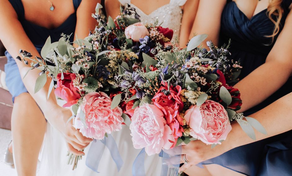 A close-up view of a floral arrangement featuring large, soft pink peonies and deep red roses, complemented by sprigs of purple lavender, white baby's breath, and silvery eucalyptus leaves. The bouque